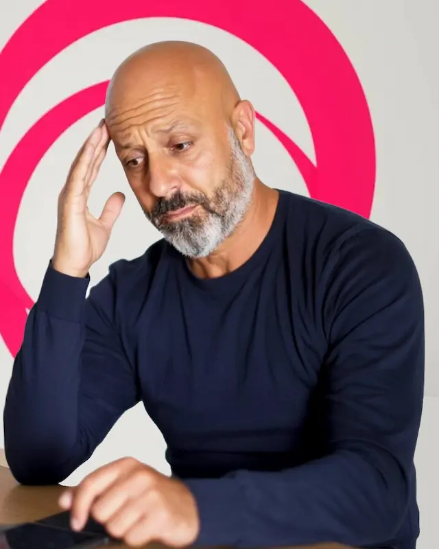 Homme concentré assis à un bureau, lisant un document avec une expression pensive, sur fond blanc décoré de cercles rose vif.